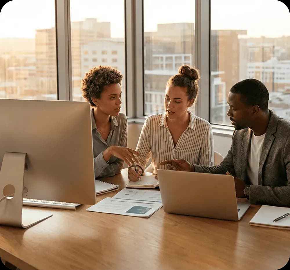 Three diverse colleagues discussing work around a desk with a computer, laptop, and documents in a modern office.