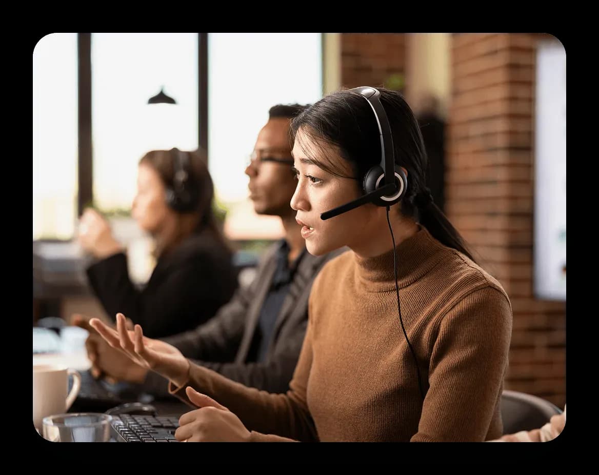 Woman with headset talking and gesturing with her hand in an office with colleagues working at computers in the background.