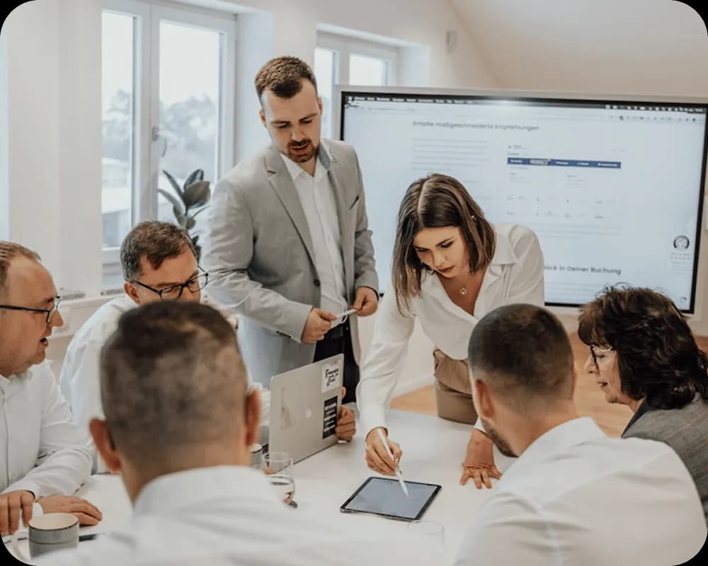 A group of six professionals having a meeting in an office, with a woman pointing at a tablet and a man standing nearby.